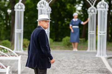 Elderly man in a hat walks towards a garden gazebo while a woman stands in the background on a sunny day