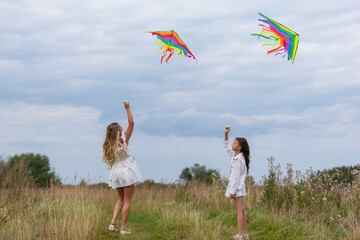 Two girls fly colorful kites in a grassy field under a cloudy sky during a breezy afternoon