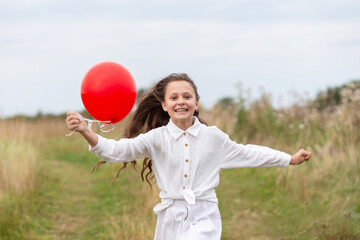 Young girl runs happily through a grassy field while holding a red balloon in her hand during a cloudy day