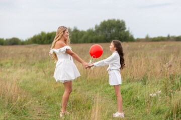 Two girls in white dresses hold hands in a grassy field while playing with a red balloon on a cloudy day