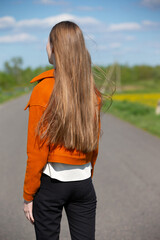 A young woman stands on a road, basking in the sunlight with a serene expression. She wears an orange jacket and black pants, surrounded by greenery and yellow flowers.