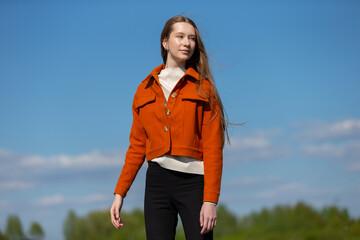Young woman in stylish orange jacket and black pants stands confidently outdoors under a clear blue sky