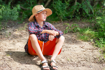 Young boy sitting on the ground in a forest, wearing a straw hat and casual clothes during a sunny day