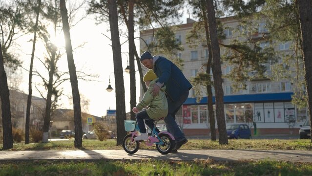 father teaches little kid to ride bike on road in city park, child spins pedals and wheels, happy family, dad helps the girl to ride daughter, play in autumn park, parent and baby have fun together.