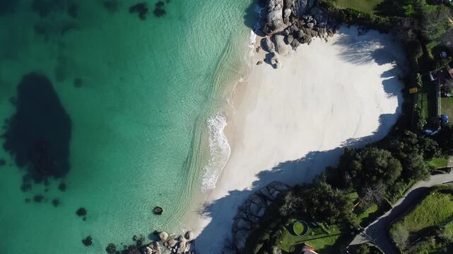 Aerial drone footage capturing the shoreline of Pintens, Galicia, Spain, with crystal-clear turquoise waters meeting the pristine white sand beach, showcasing intricate sand and wave patterns  
