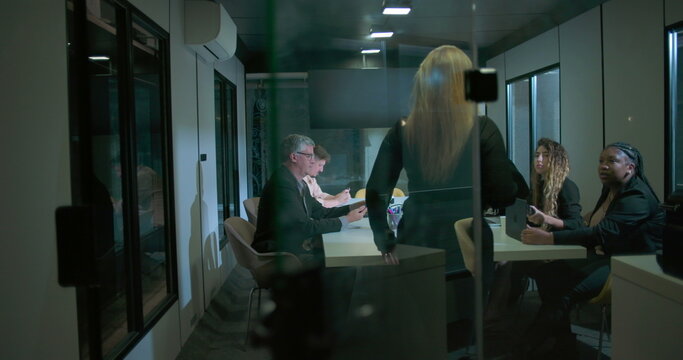 Back view of woman entering business meeting as team members look up in modern glass conference room during evening work session