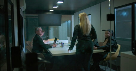 Back view of woman entering business meeting as team members look up in modern glass conference room during evening work session