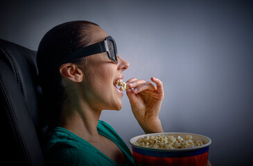 A woman wearing 3D glasses sits in a dark room, intently watching a screen while vigorously eating popcorn from a large red and blue bucket.