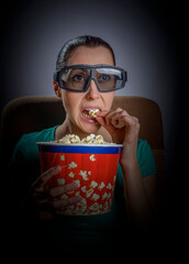 A woman wearing 3D glasses sits in a dark room, intently watching a screen while vigorously eating popcorn from a large red and blue bucket.
