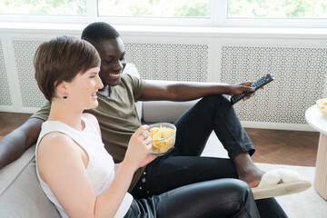 Young african american man and causasian woman watching TV and eating chips on the floor