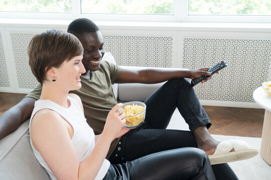Young african american man and causasian woman watching TV and eating chips on the floor