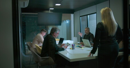 Back view of woman entering business meeting as team members look up in modern glass conference room during evening work session