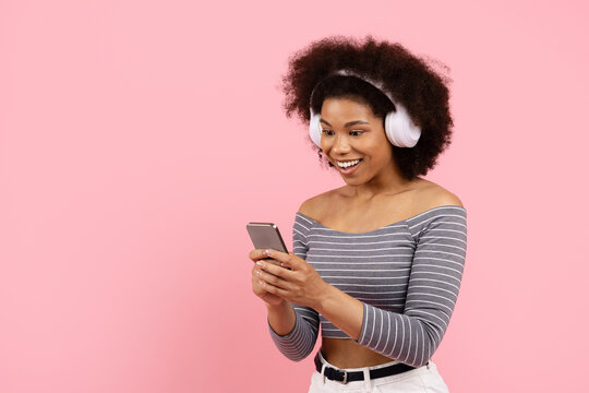 Excited African American woman wearing headphones and looking at smartphone, smiling with surprise against pink background. Concept of music, message, or good news