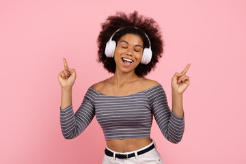 Joyful African American woman with headphones dancing and listening to music on pink background. Concept of happiness, rhythm, and self-expression