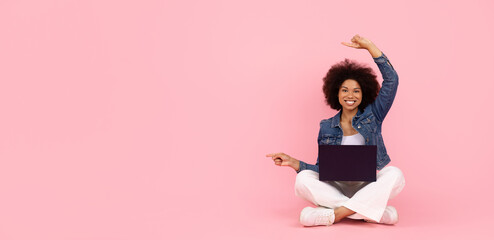 Smiling African American woman sitting with laptop and pointing at copy space on pink background. Concept of online creativity and multitasking, panorama