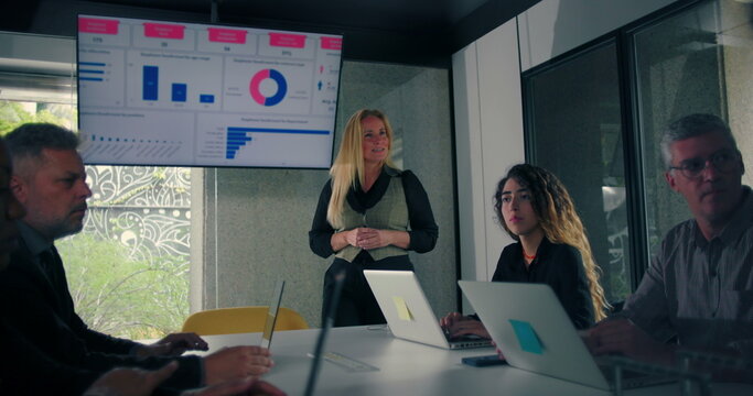 Blonde businesswoman delivering presentation to engaged colleagues seated around conference table with data screen in modern office - Powered by Adobe