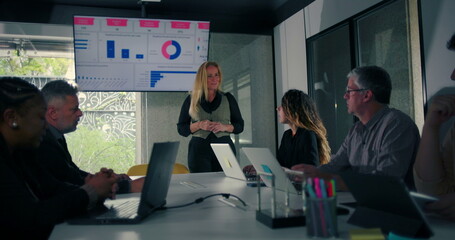 Blonde businesswoman delivering presentation to engaged colleagues seated around conference table with data screen in modern office