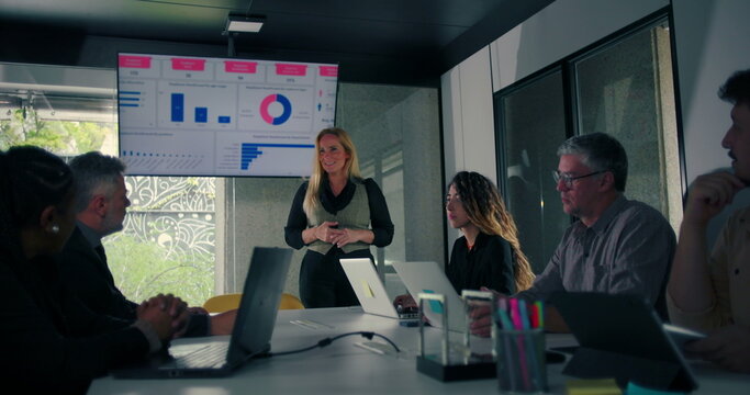 Blonde businesswoman delivering presentation to engaged colleagues seated around conference table with data screen in modern office