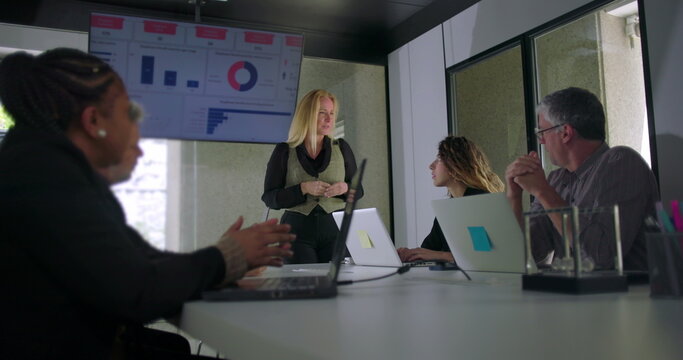 Blonde businesswoman giving presentation in front of data screen as coworkers watch and interact in professional modern office setting - Powered by Adobe