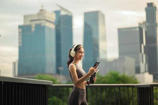 Asian woman wearing headphones standing outdoors in an urban city environment, happily checking her smartphone while holding a water bottle after a workout session.