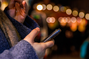 A closeup of hands holding a smartphone against a blurred background of warm festive lights at a...