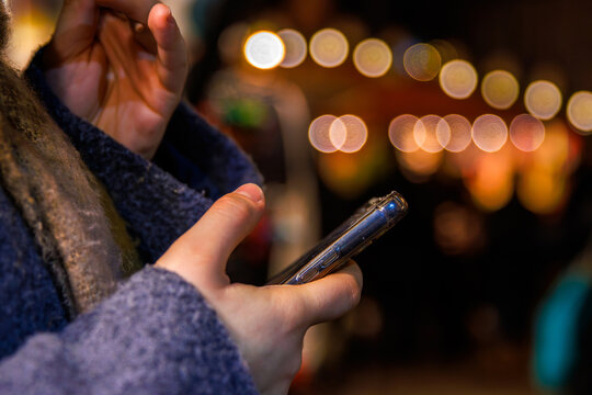A closeup of hands holding a smartphone against a blurred background of warm festive lights at a night holiday market. Selective focus