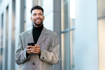 Smiling man in suit with smartphone on city street, low angle view. Space for text