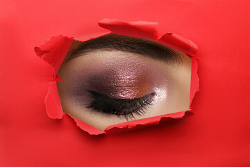 Young woman with evening makeup, closeup. View on eye through torn hole in red paper