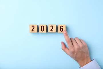 New 2026 year. Man putting cube with number 6 on light blue background, top view