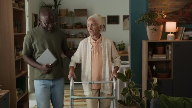 Medium shot of Black man as social worker assisting white haired senior woman using walker and slowly walking towards camera at home, copy space