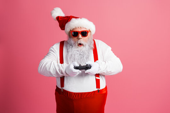 Santa Claus in red suit with suspenders and beard plays a game controller on a pink background for Christmas celebration and holiday fun