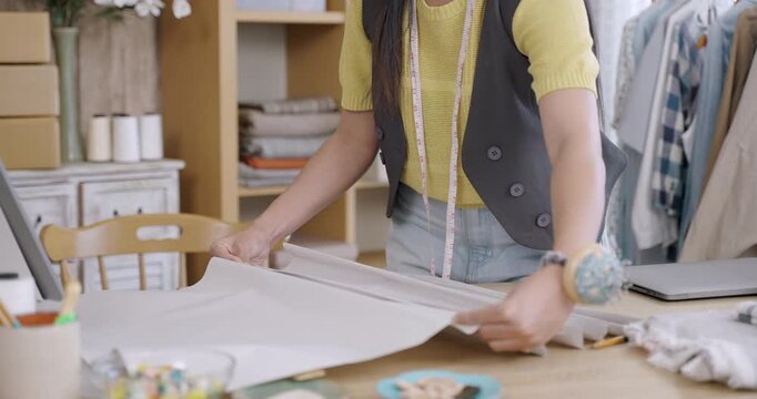Young asian female fashion designer working on paper pattern layout for handmade garment while arranging pieces on table inside tailoring studio filled with tools and textile materials