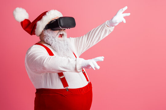 Santa in a virtual reality headset reaches out in a festive pose wearing red suspenders and a hat in a pink studio with a gleaming holiday vibe