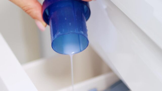 Hand pouring liquid laundry detergent or fabric softener into washing machine &mdash; clothes washing and home cleaning routine concept	