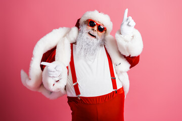 Santa in red suit with white fur and sunglass points up while smiling against a pink background...