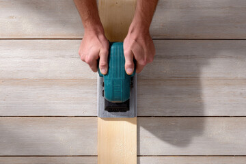 Overhead close up of hands guiding cordless planer across wooden plank creating smooth surface with wood shavings