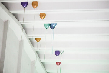 Colorful metallic balloons tied to thin strings, nestled between the white beams of an industrial ceiling.