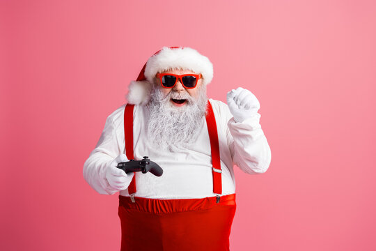 Santa in red suspenders with white beard on pink background celebrates Christmas wearing hat coat and funny sunglasses