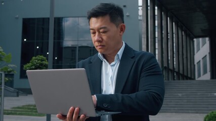 Asian middle-aged businessman typing laptop in city Korean business man serious browsing wireless computer in downtown internet virtual job work task startup project Chinese Japanese male entrepreneur - Powered by Adobe