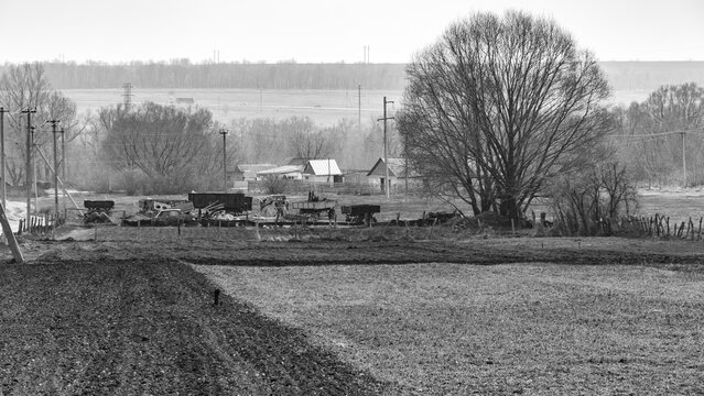 Rural landscape with fields, old farm equipment, utility poles, and distant houses on a misty day, captured in black and white - Powered by Adobe