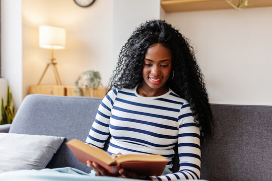 Young black woman enjoying her leisure time reading a book while relaxing on a comfortable sofa in her modern living room