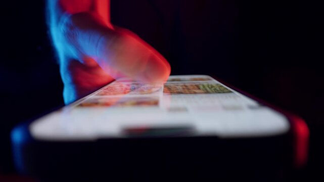 Finger Scrolls Food Images on Smartphone Screen with Intense Red and Blue Lighting, Low Angle Emphasizing How the Whole Life is in the Smartphone Through Visual Consumption