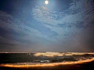 Under the moonlit sky, where sea waves kiss the shore and clouds drift over a tranquil blue, the night reflects a serene dance of light and water.