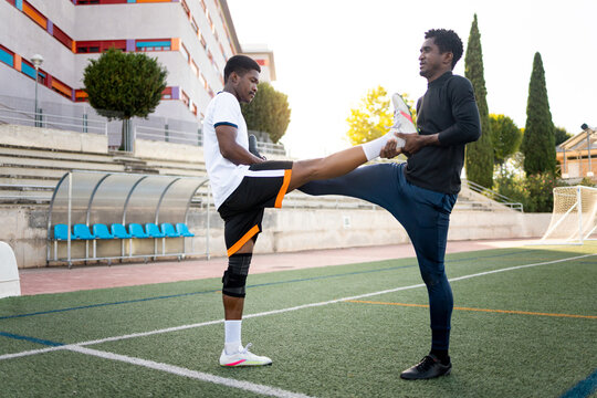 Two dark-skinned soccer players are stretching their legs together on a grass soccer field, wearing white uniforms. Grass soccer concept. African boys training.