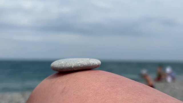 Horizontal video close up. Male hand builds small rock pyramid on female knee. Blue sky. Sun, sea waves, coastline. Concept of rest on pebble beach, calm, yoga, travel, ocean shore zen, tourism, relax