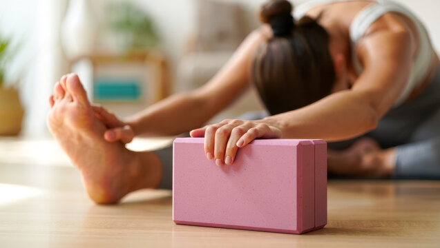 Close-up of a woman stretching forward in a calm yoga pose, using a pink support block for balance, with soft natural light and warm tones creating a peaceful, mindful home practice atmosphere