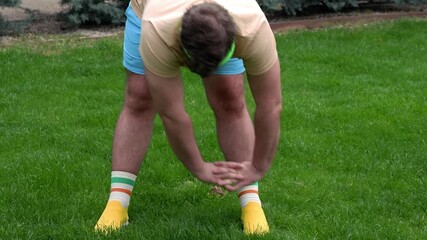 Horizontal street video. 60 fps. Mature fat white man with beard, big belly, blue shorts, beige t-shirt, green headband, yellow sports sneakers doing bends in city garden at summer day. Fitness day