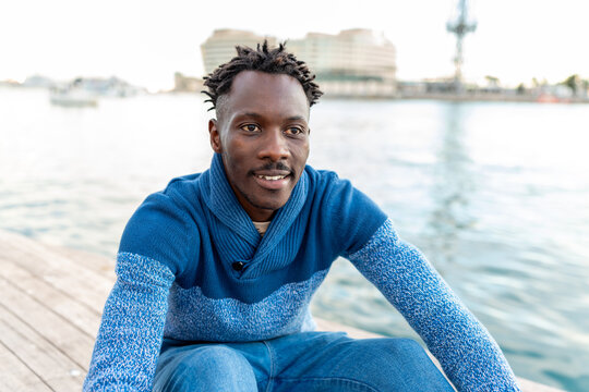 African man sitting on a wooden pier, enjoying his vacation in Barcelona and smiling
