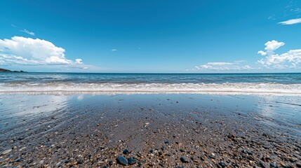 A serene view of a pebble beach with the ocean waves gently lapping the shore under a bright blue sky dotted with fluffy white clouds.