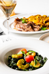 Two plates of elegant Italian cuisine: black tagliatelle with shrimp/tomatoes (foreground), yellow pasta with steak (background), served with sparkling wine in a bright setting